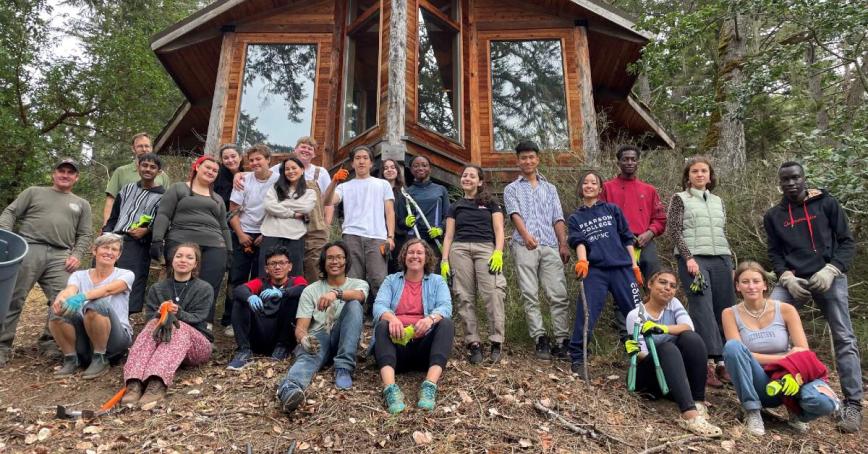 Group of smiling students from Pearson College UWC standing in front of a cabin in the forest.