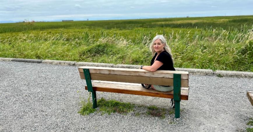 Lesley Pollard seated on a bench, turning to look at the camera with a meadow behind her.