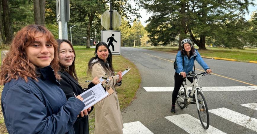 Three students standing in front of a crosswalk with clipboards and a cyclist stopped in the background.