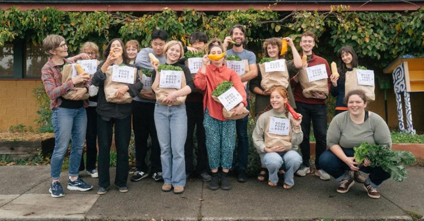 Group of people holding Fernwood Neighbourhood House Good Food bags. They are holding up different fruits and vegetables and smiling.