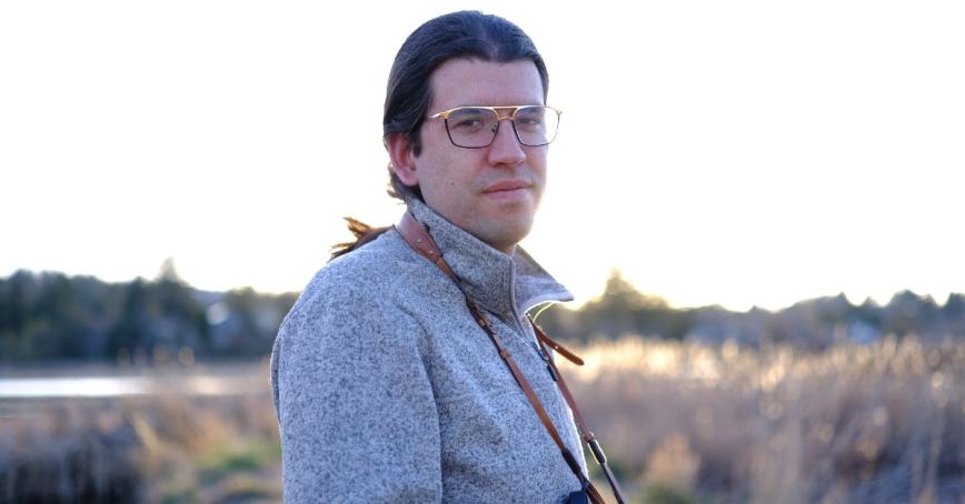 Jeff Nicholls is looking at the camera with trees, water, and low brush vegetation in the background.