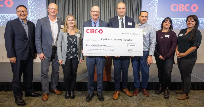 Nelson Chan, Matt Gillberg, Jacky Weber, Philip Steenkamp, Spencer Walker, Russell Johnston, Cherylanne James, Shelby Blackjack stand in a line. Philip and Spencer hold a large cheque for $100,000 from CIBC to Royal Roads University.
