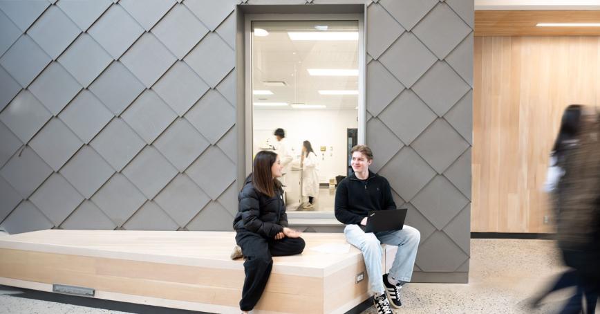 Two students sit on a wooden bench in front of a lab inside an RRU building