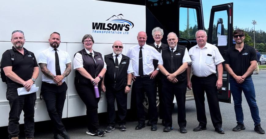 Group of smiling employees in front of a passenger bus with Wilson's logo on the side of it.
