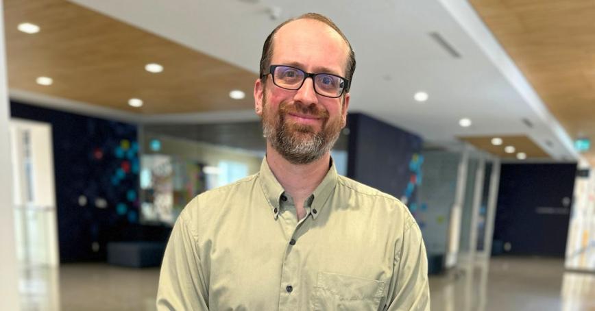 A man in glasses, poses for a headshot in a hallway.