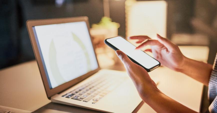 A hand holds a mobile phone, with a laptop open in front of it