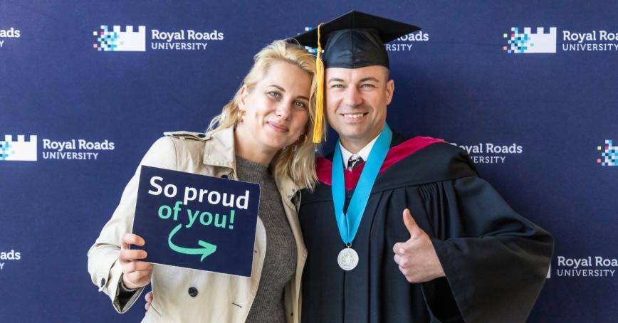 A graduate with a meal around his neck holds a thumbs up while a woman next to him holds a sign that reads, "so proud of you"