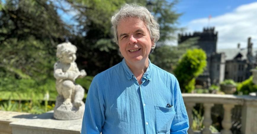 A man in a blue collared shirt smiles. A cherub statue is visible in the background.