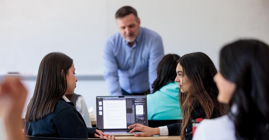 Students engaged in a classroom discussion with a teacher leaning over a desk