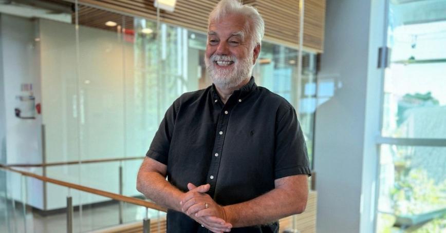 A man gestures while speaking in a building in front of a window.