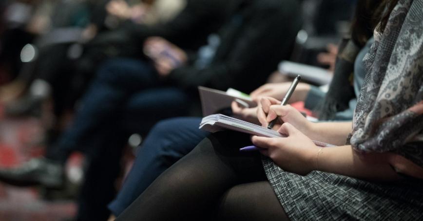 Woman holding a notebook in her lap and taking notes, her face is not seen in the image.