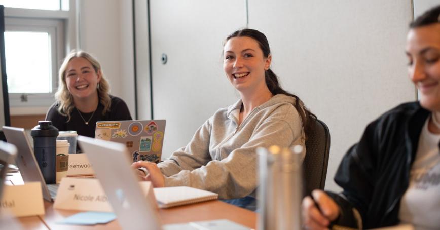 Three young women sitting at a table with laptops open and smiling.