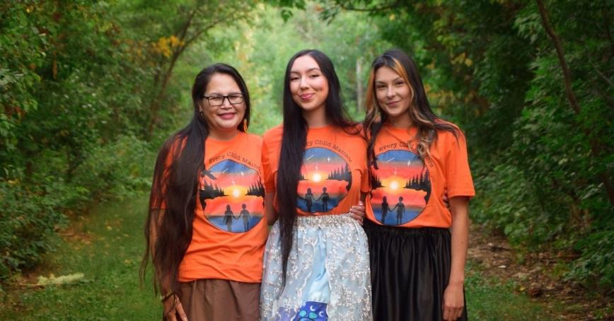 Three women, wearing orange shirts and ribbon skirts, pose in a forested area.
