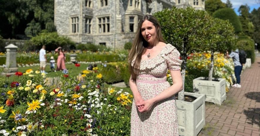 A woman poses in a colourful flower garden, with a stone castle in behind her.