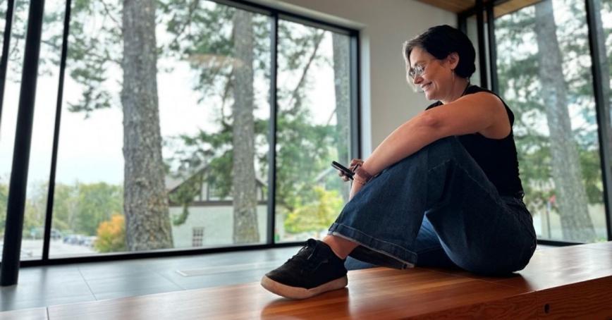 A woman scrolls on her phone in a lobby, trees can be seen out the window.