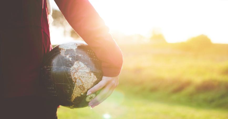 Person holding a globe by their side with trees and sunshine in the background.