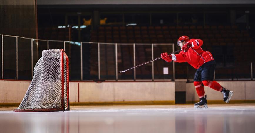 A man in hockey attire on an ice rink hitting a puck into the net. 