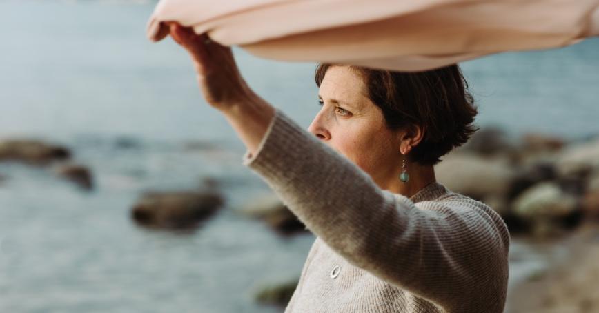 Cheryl Heykoop standing on a beach holding a pink scarf high in the wind.