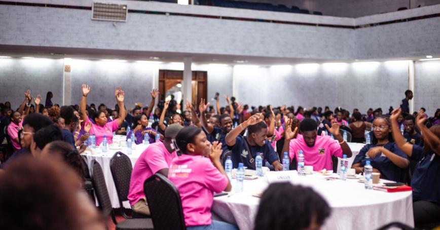 groups of people seated at round tables wearing hot pink t-shirts, their hands are raised.