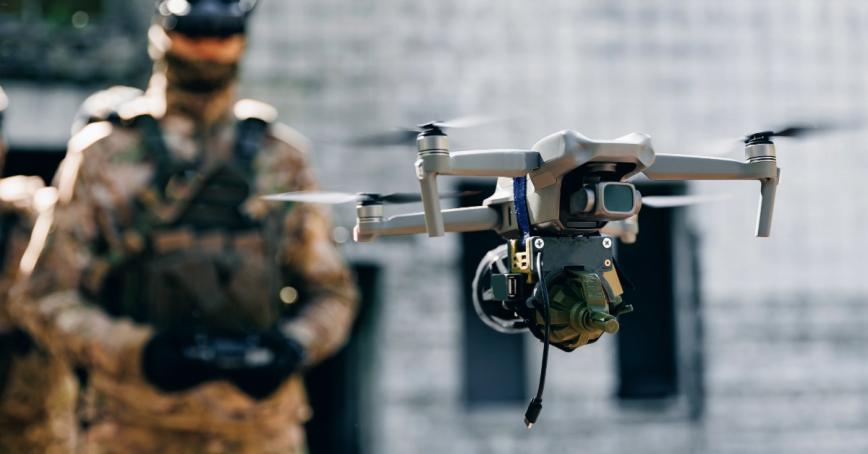 A soldier in combat fatigues using a handheld console to fly a drone, featured in the foreground