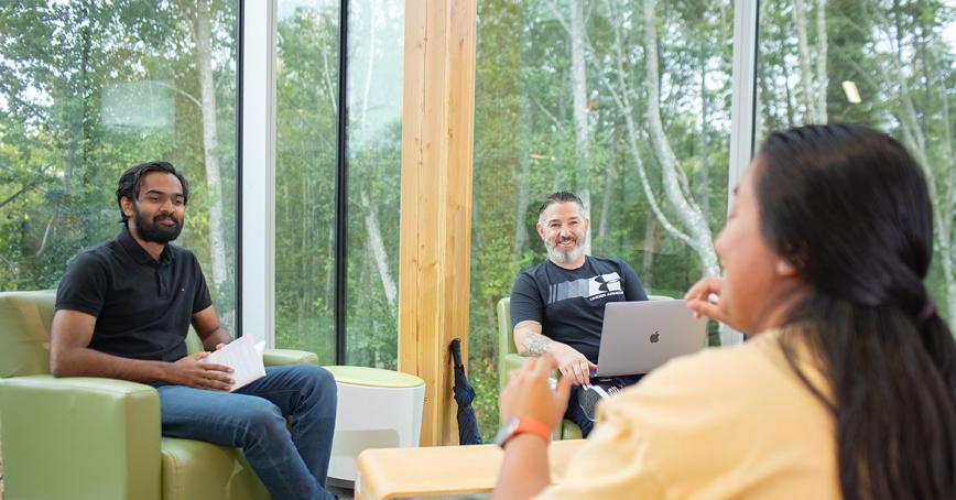 RRU students sit across from each other in an engaged discussion, surrounded by windows and outdoor foliage.