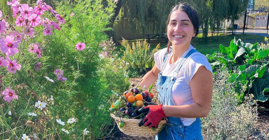 Emily Mulroney holding a basket of vegetables standing by tall flowers in The Farm at RRU
