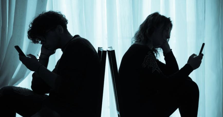 Two teenagers in a dark room, back to back, staring at their smartphones