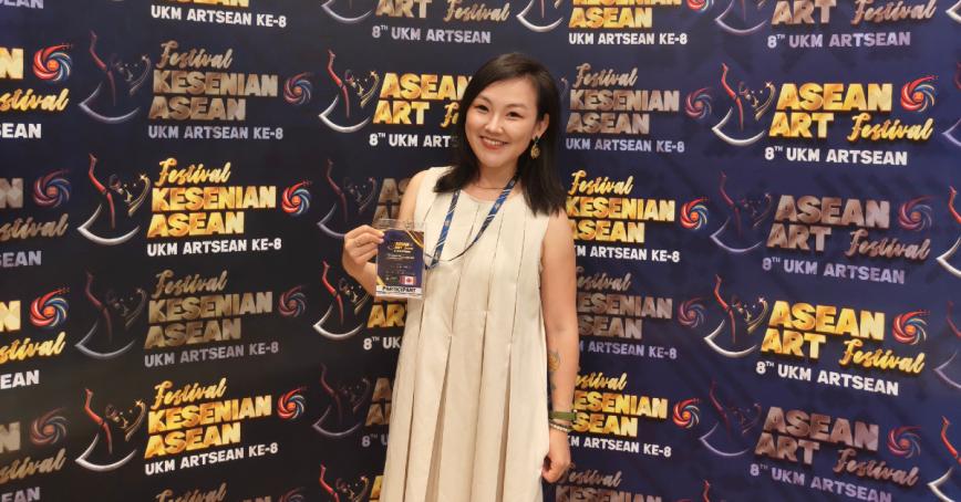 Shuwen Tian holding up her delegate badge and standing in front of ASEAN ART Festival 8th UKM Artsean backdrop