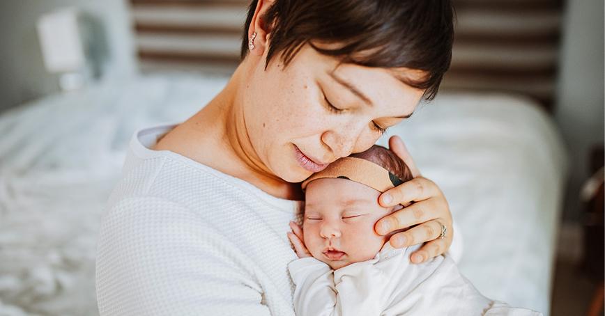 A mother gently holding and cuddling a sleeping newborn baby, resting against their chest in a calm indoor setting.