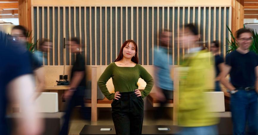 A photo of a woman looking at the camera. The people around her appear blurred by their movement.