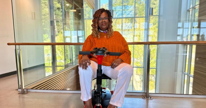 A woman smiles at the camera. She is indoors, in front of a glass wall. She's sitting in a red mobility scooter.