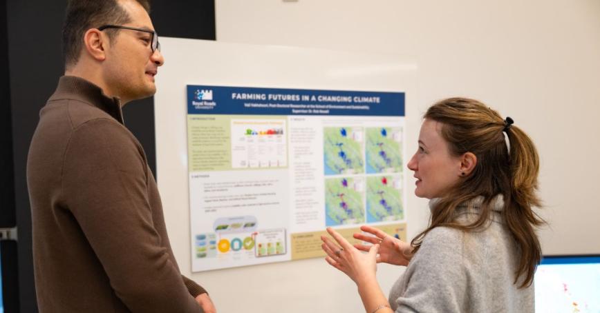 A woman gestures while speaking to a man, a poster board behind them.
