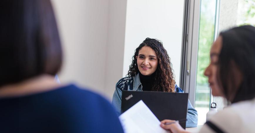 RRU students sit around a table by a large window with their laptops and engage in conversation