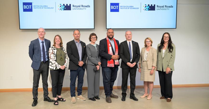 A group of BCIT and RRU staff stand aligned under screens showing both institutions' logos