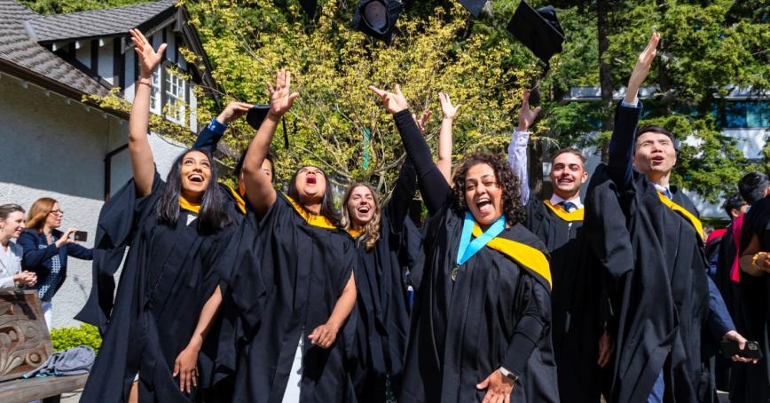 A group of graduates in convocation robes with yellow sashes throw their grad caps in the air and smile