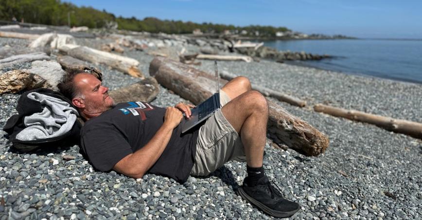 A man lays on a rocky beach, surrounded by driftwood. He's laying down, working on his laptop.