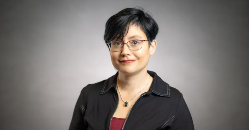 A studio head shot of a woman with a gentle smile in front of a grey screen. She has short, cropped hair.