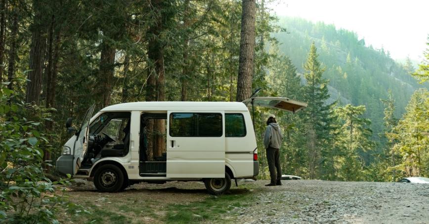 A camper stands beside their van in a forested area