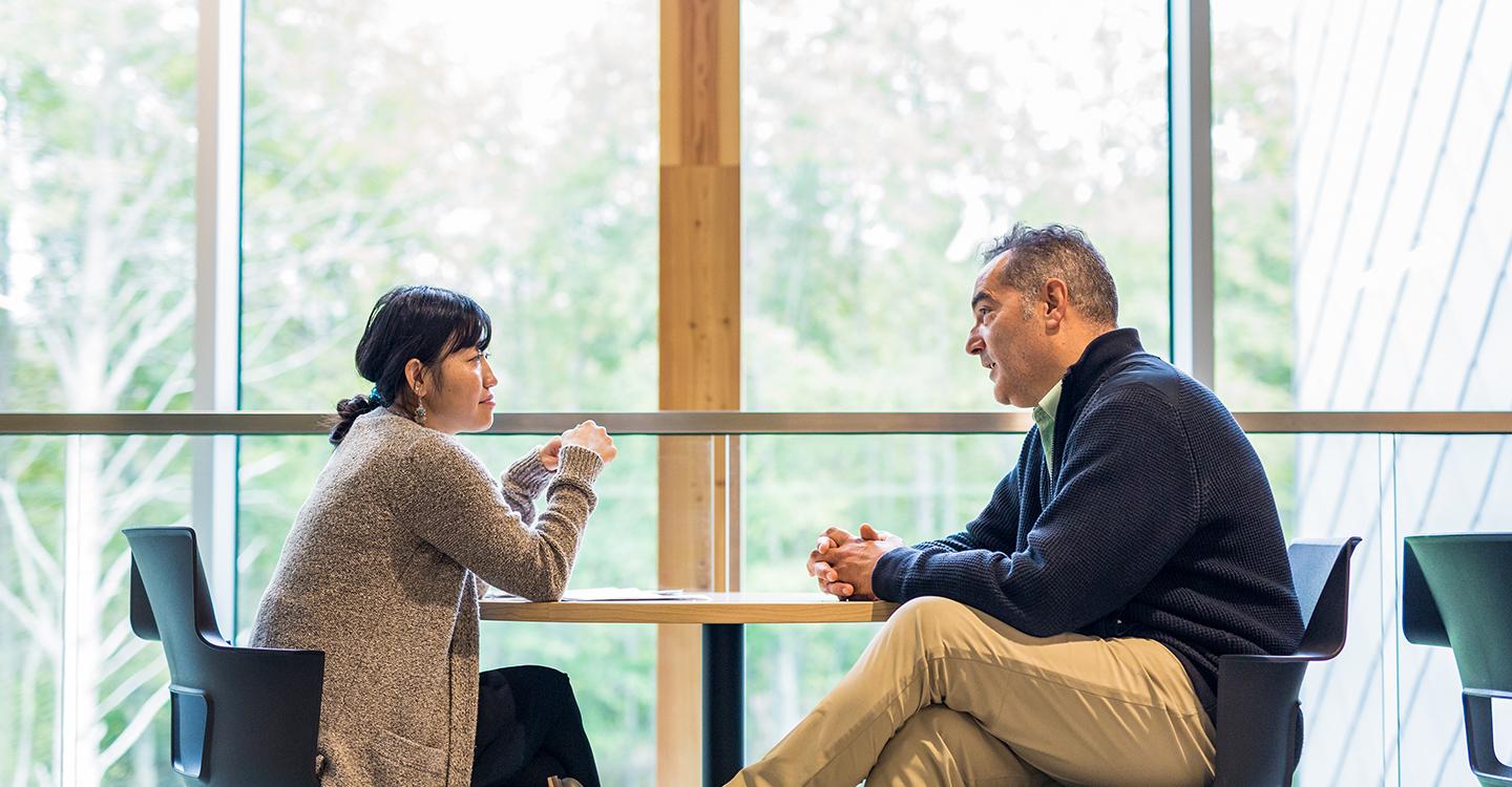 An international student sits with an advisor at a table in front of a large window in the Sherman Jen Building.