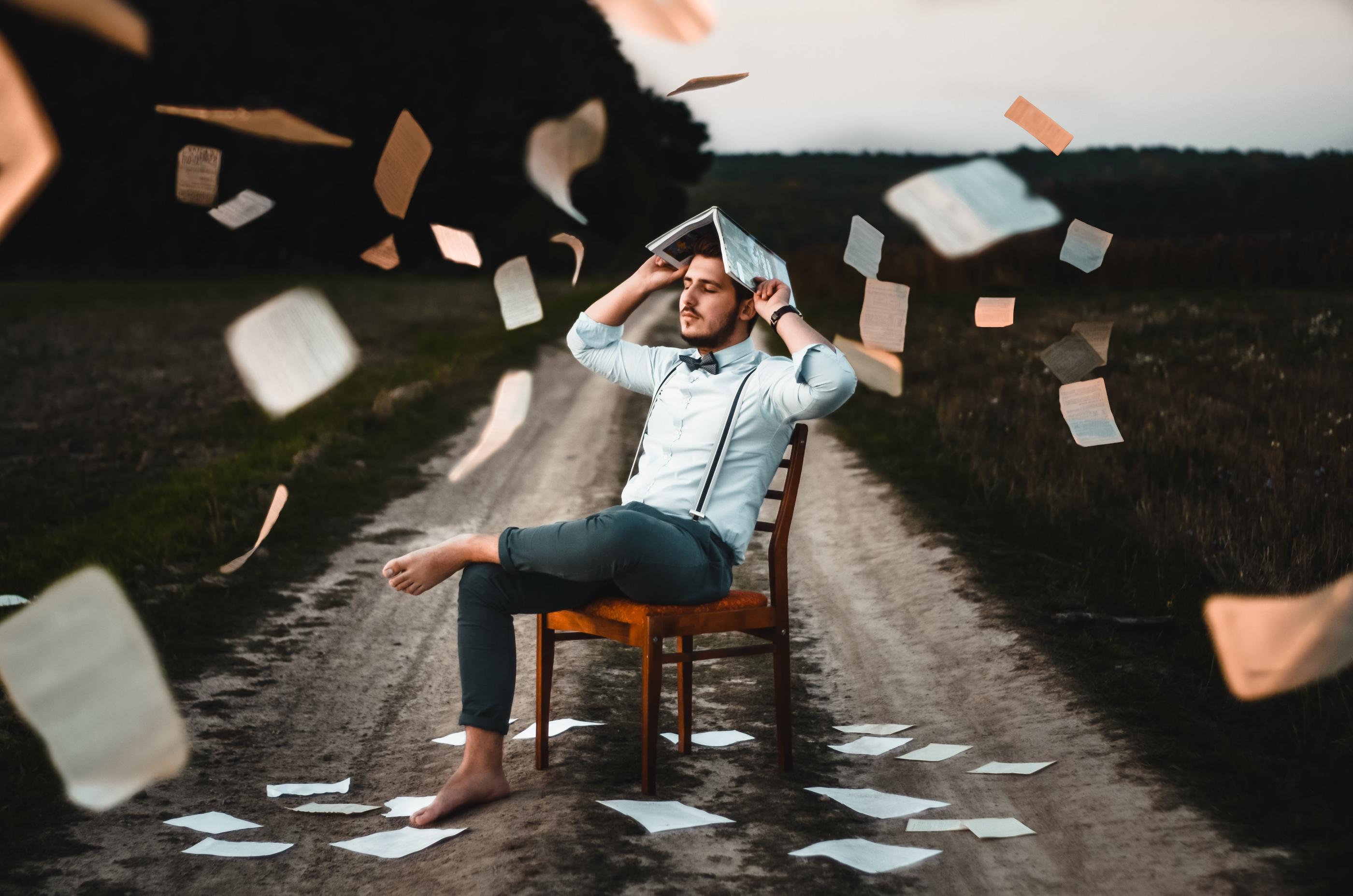 A person sitting on a chair outside on a dirt road, eyes closed, with an open book on the end with pages flying around