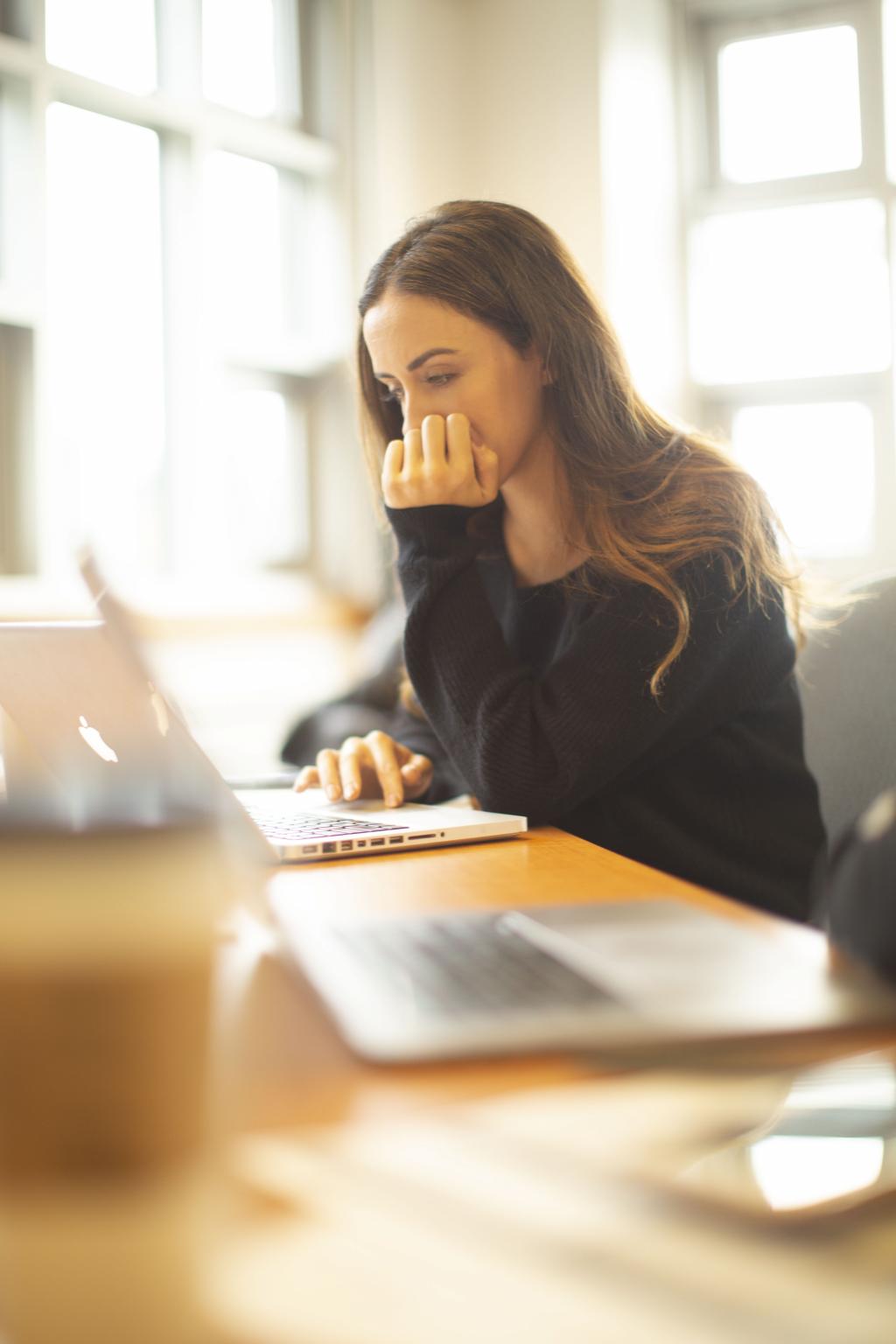 A student concentrates on the screen of her laptop while sitting at a table in front of classroom windows.