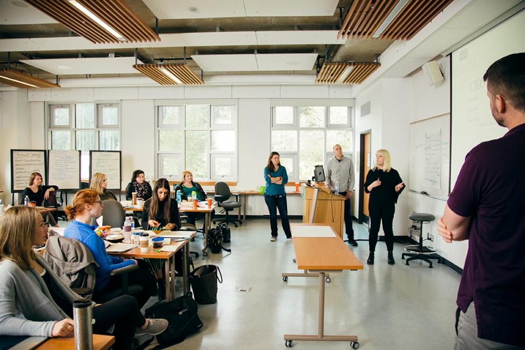 Students sit at tables in a classroom, watching as a group of classmates give a presentation next to a whiteboard.