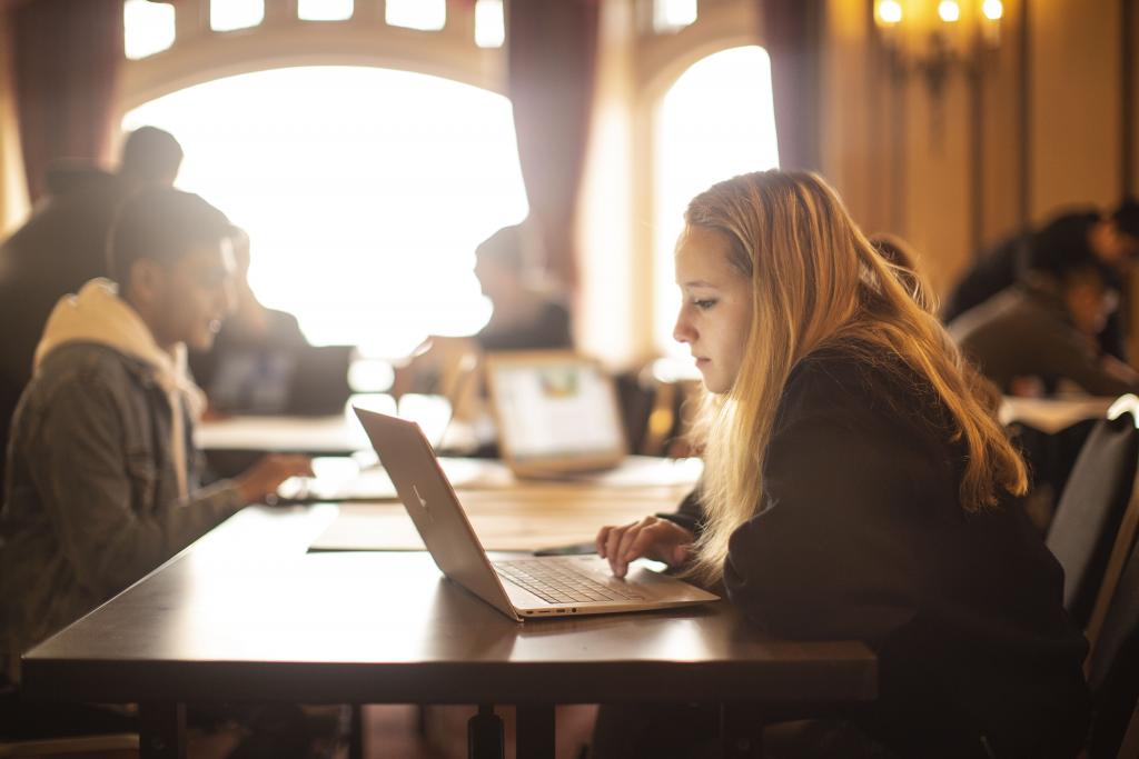 A student with long blonde hair on sits at a table on a laptop with other students in the background.