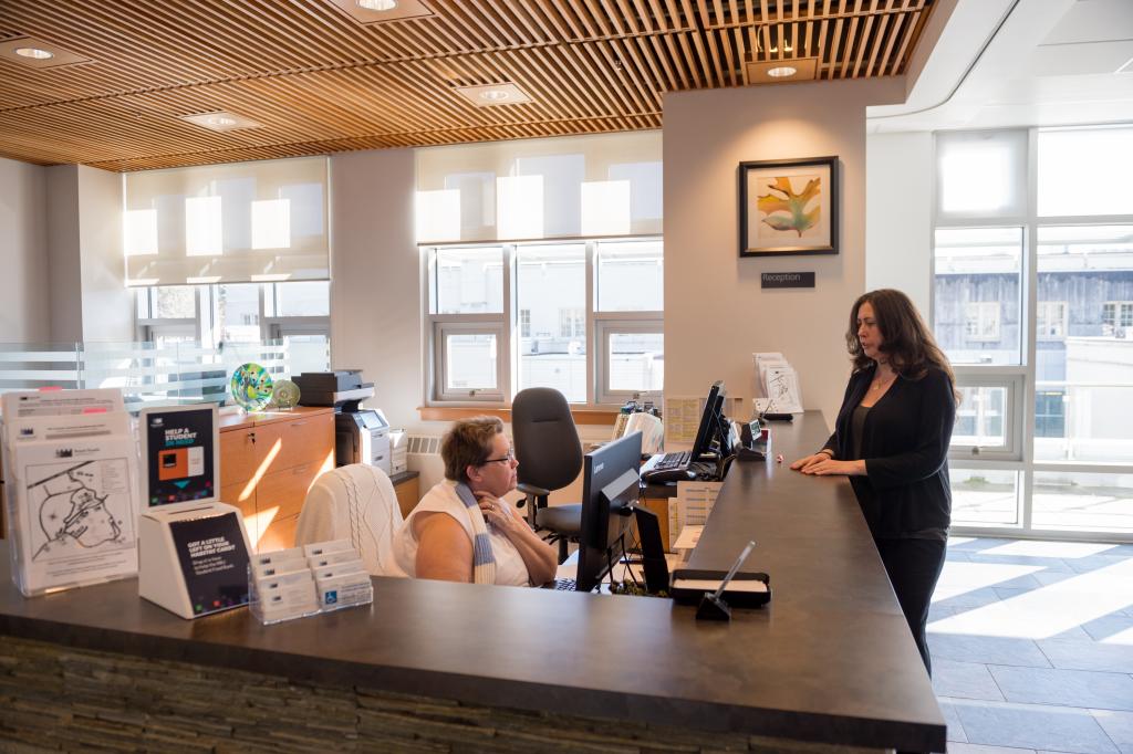 Person sitting at a reception desk speaks with someone standing at the front counter