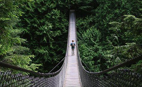 A person with a backpack crosses a long bridge into green trees. 