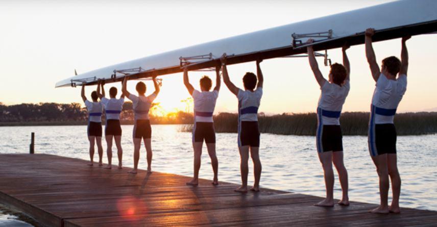 A rowing team holds their boat over their heads while standing on a dock as the sun sets.