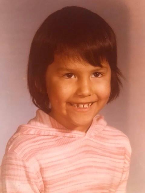 Corinne Lewis-Coutre as a young child. Her pink hoody matches a soft pink background. She is smiling and has cute short hair.