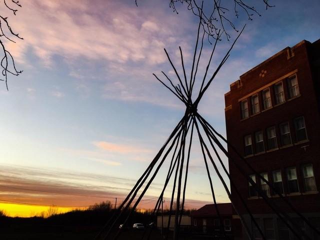 The residential school Corinne Lewis-Couture's mother attended. You can just see the building to the right. In front, the structure of a teepee is a shadow against a soft sunset.