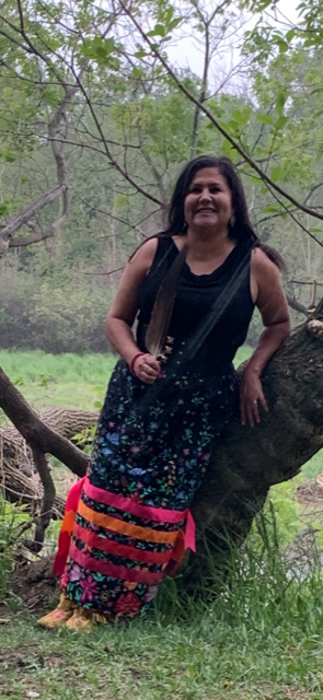 Corrine Lewis-Couture as an adult leans against a tree. She is wearing a beautiful black dress with ribbons of colour around the bottom.