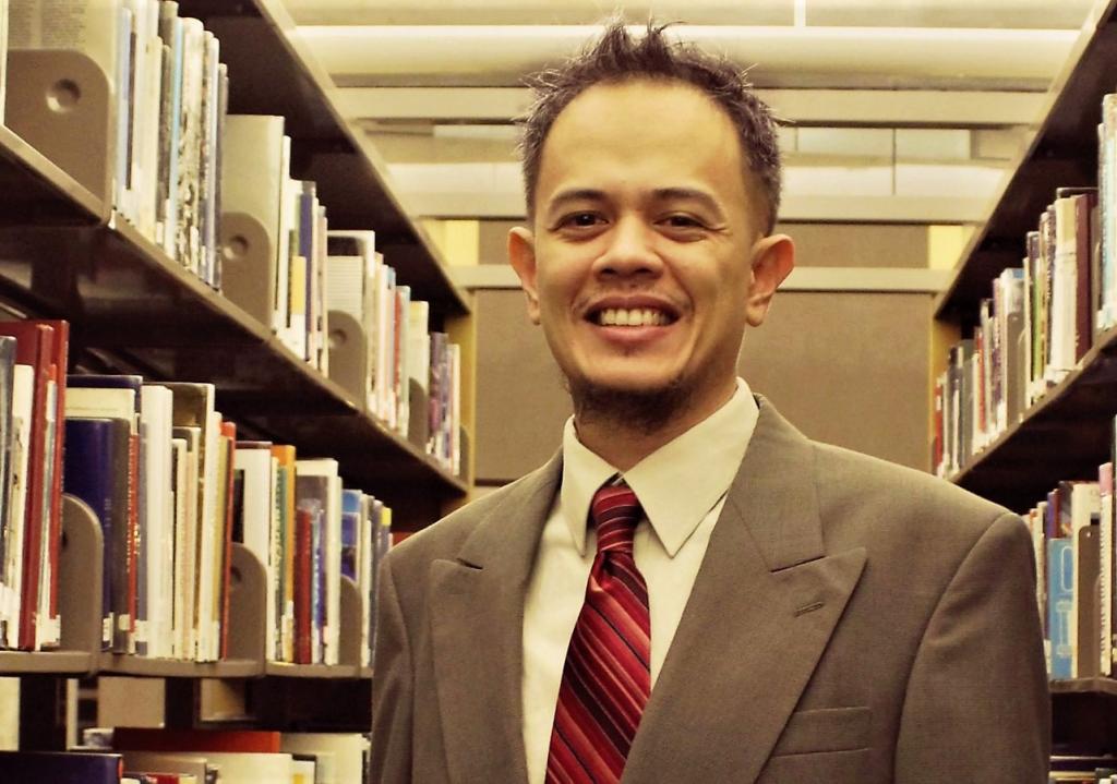 Man in suit and tie smiles from between stacks of books in library